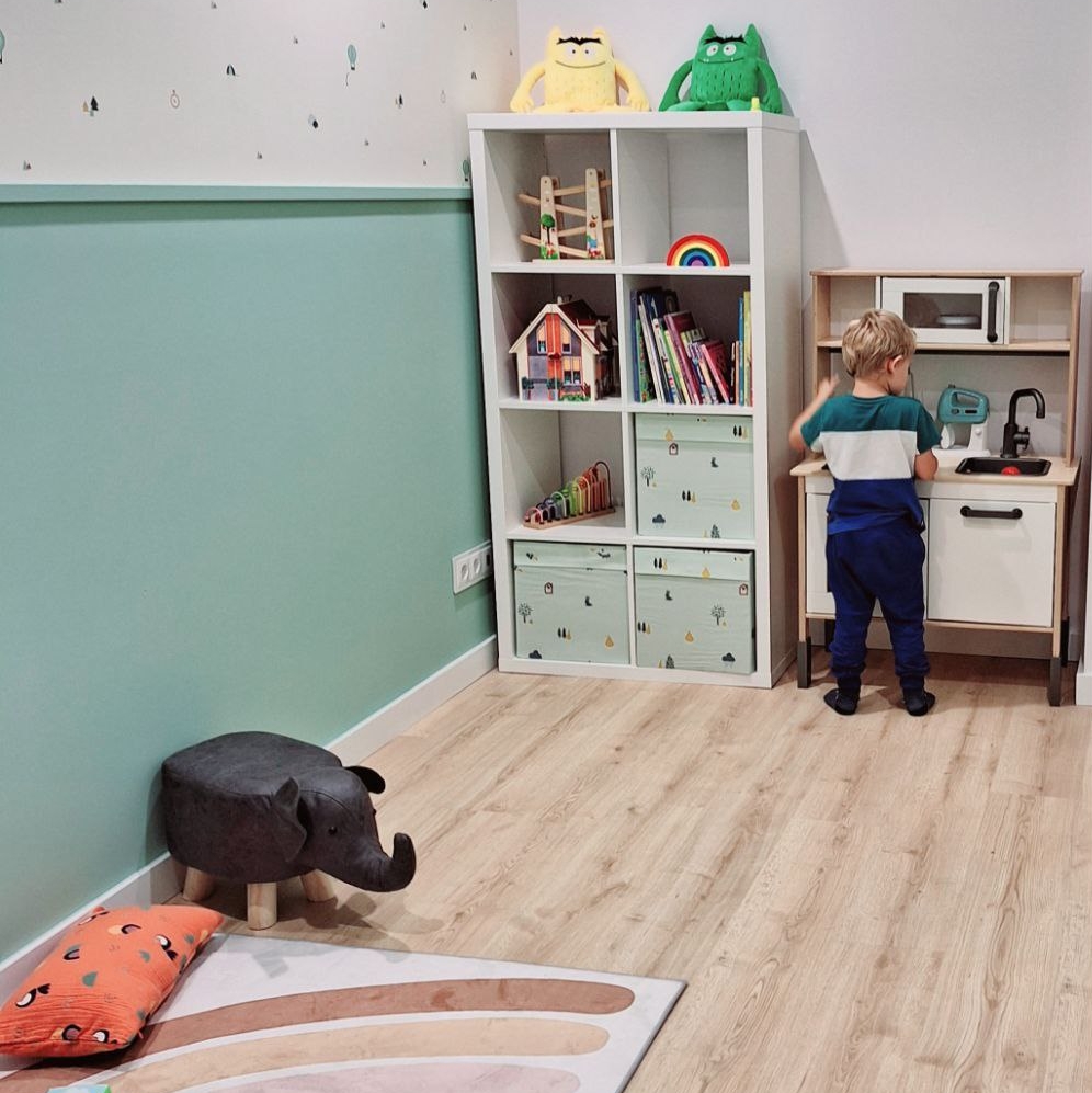 A cupboard with books and toys. A child is playing in front of a toy kitchen. A carpet with toys and a pillow. A small table with two chairs for children.